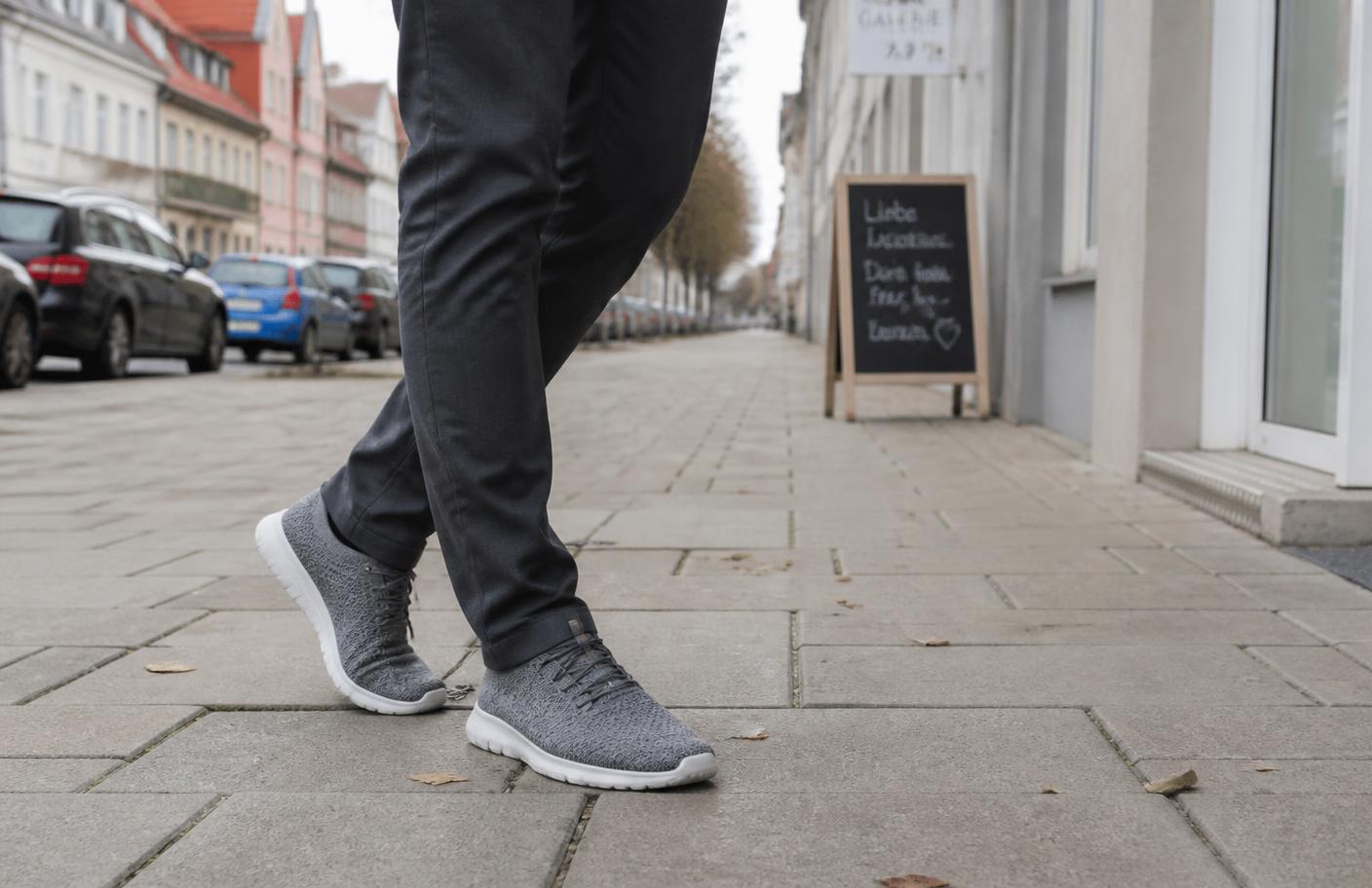 Person in dunkler Hose und grauen Strickschuhen geht über einen gepflasterten Gehweg in einer deutschen Innenstadt